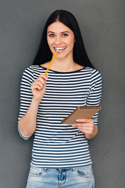 Waiting for inspiration. Attractive young woman holding note pad and carrying pencil in mouth while standing