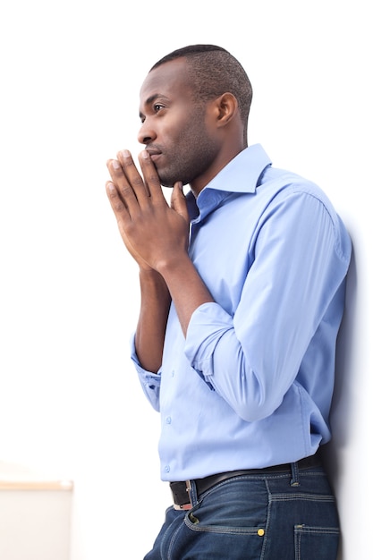 Waiting for inspiration. Side view of handsome young black man keeping his hands clasped and looking away while leaning at the wall