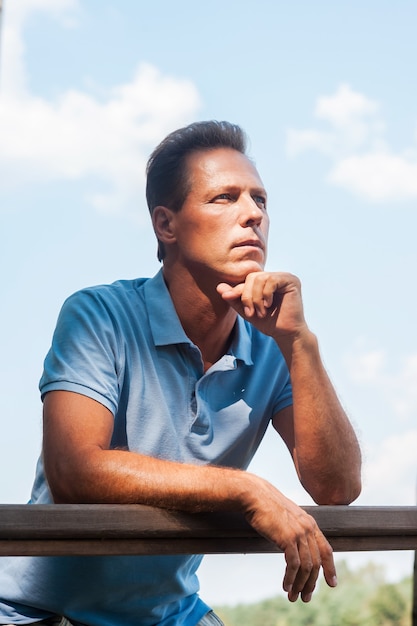 Waiting for inspiration. Thoughtful mature man holding hand on chin and looking away while leaning at the wooden fence