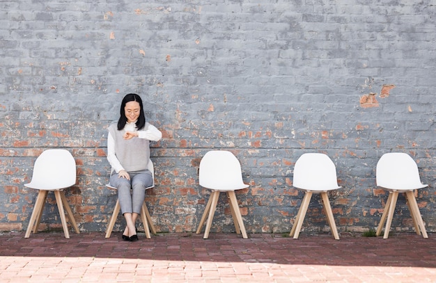 Waiting interview and woman check time for recruitment job opportunity and human resources feedback Asian person alone on chair looking at watch for career news feedback and appointment schedule
