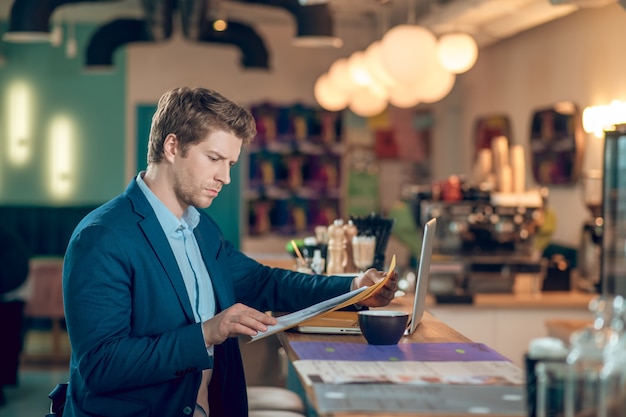 In attesa. giovane serio in giacca e cravatta che legge il documento nella cartella mentre è seduto davanti al laptop e al caffè in un caffè illuminato