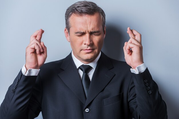 Waiting for special moment. Portrait of mature man in formalwear keeping fingers crossed and eyes closed while standing against grey background