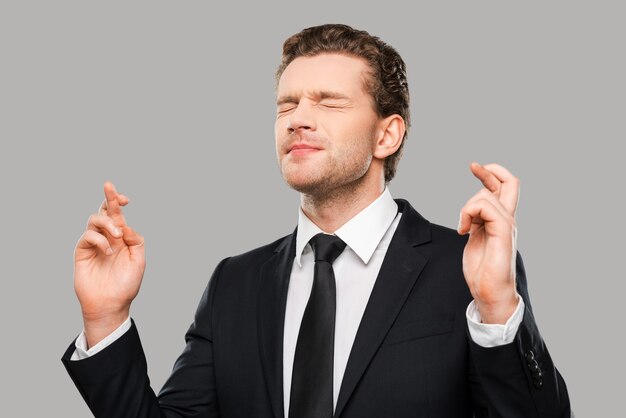 Waiting for special moment. Portrait of young man in formalwear keeping fingers crossed and eyes closed while standing against grey background