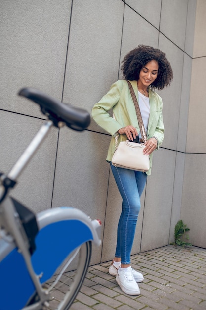 Waiting. Woman in jeans and jacket standing near the bike and taking the phone out of the bag