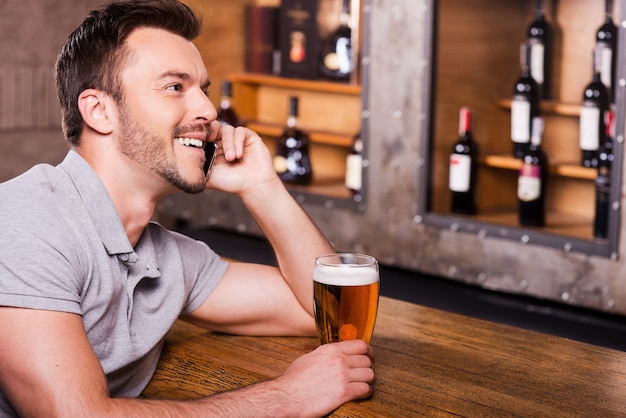 あなたを待っています、仲間！ビールとグラスを保持し、バーカウンターに座って携帯電話で話している陽気な青年の側面図