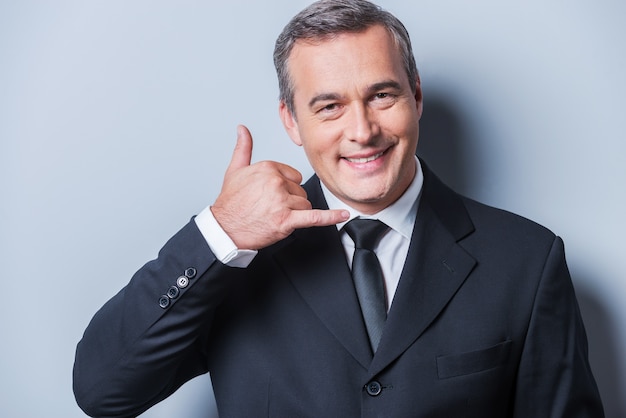 Waiting for your call. Confident mature man in formalwear gesturing mobile phone near his face and smiling while standing against grey background