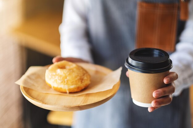 Una cameriera che tiene e serve un pezzo di ciambella fatta in casa in un vassoio di legno e una tazza di caffè di carta