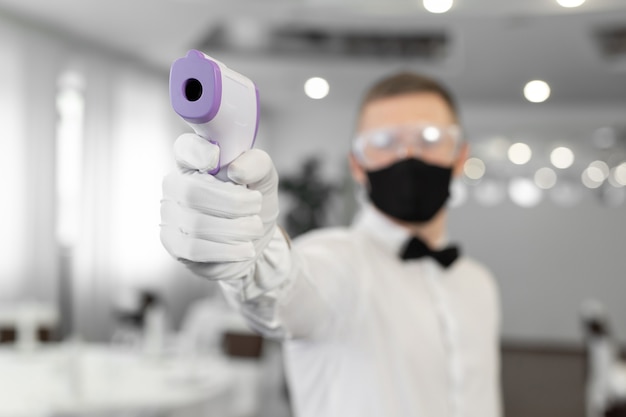 Waitress man wearing a face mask and holding an infrared thermometer on her forehead to check the body temperature for symptoms of the virus in customers