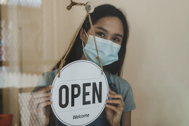 Waitress staff wearing protection face mask turning open sign\
board reopen after coronavirus quarantine is over in cafe coffee\
shop ready to service reopen cafe restaurant food and drink\
concept