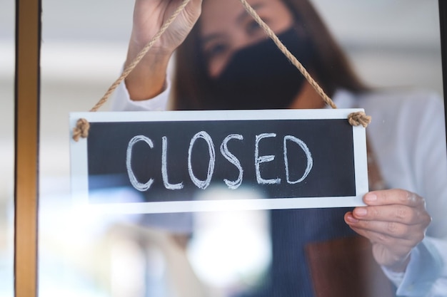 A waitress wearing protective face mask hanging closed sign on front door due to coronavirus pandemic effect