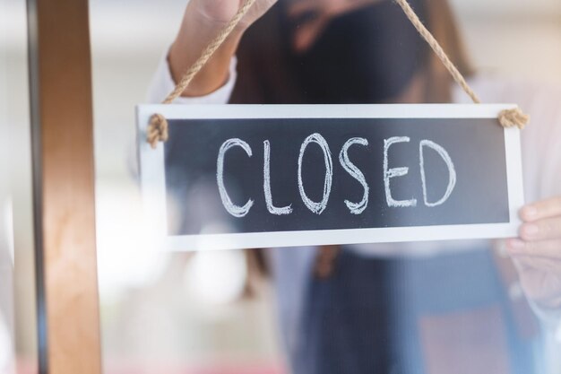 A waitress wearing protective face mask hanging closed sign on front door due to coronavirus pandemic effect