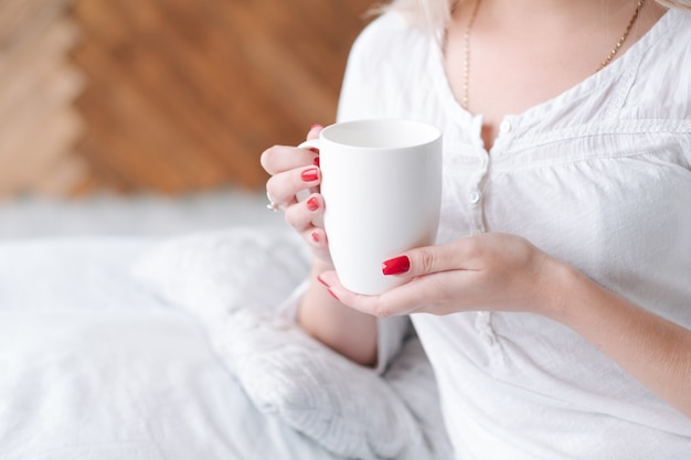 目覚めの飲み物。熱い朝の飲み物の白いカップを保持しているベッドの女性。