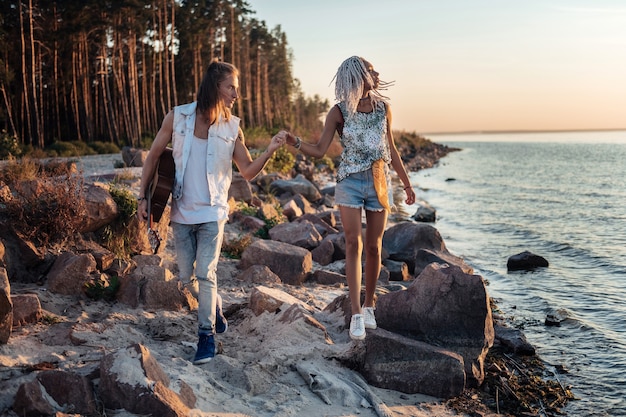 Walk along beach. Caring blonde-haired man holding hand of his girlfriend while walking along the rocky beach