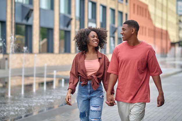 walk. young african american woman and attractive man smiling looking at each other hand in hand walking down street against background of building and fountain