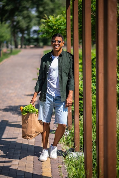 歩く。夏の日に公園でカメラに微笑んで立っている食品のバッグとサングラスの浅黒い肌の若者