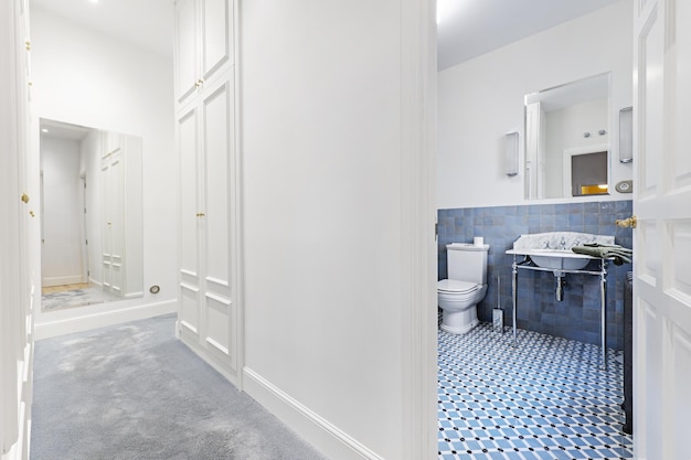 Гардеробная с зеркалом на стене, бледно-серым ковровым покрытием и выходом в ванную комнату с винтажной мебелью в голубых тонах.