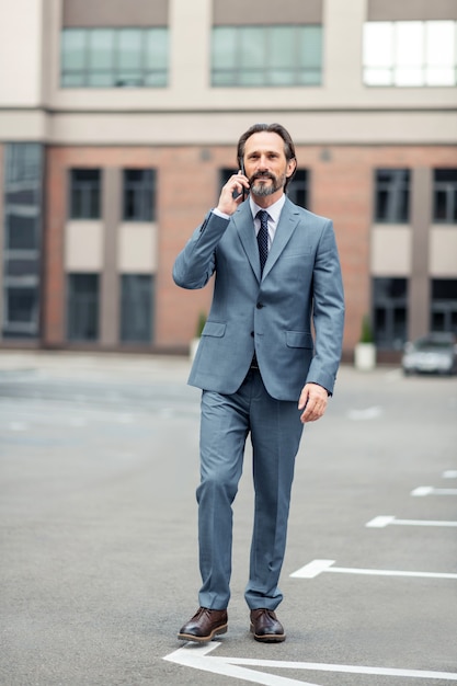 Walking by foot. Grey-haired mature man wearing grey suit calling colleague while walking by foot