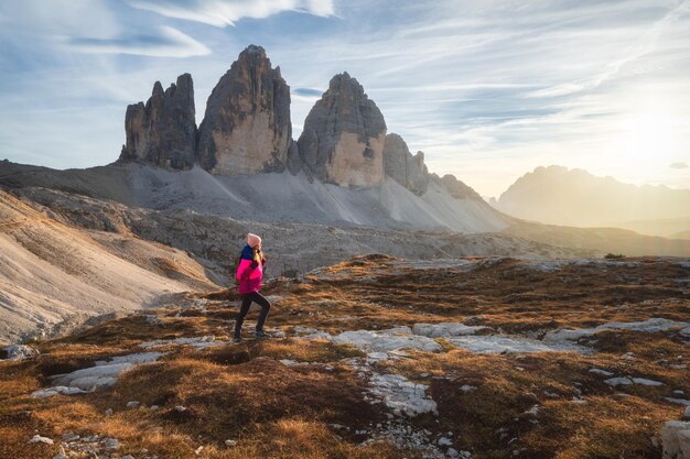 Walking girl with backpack on the trail in mountains at sunset in autumn Tre Cime Dolomites Italy Beautiful landscape with young woman high rocks path stones orange grass sky in fall Hiking