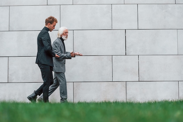Walking on grass near wall Young guy with senior man in elegant clothes is outdoors together Conception of business