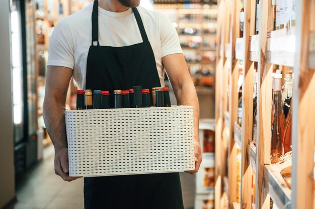 Foto cammina e tiene un mucchio di bottiglie proprietario di un negozio di vino in camicia bianca e grembiule nero