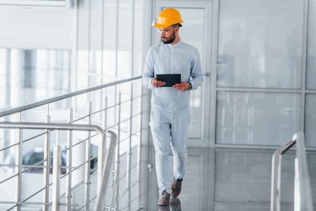 Walking near silver colored railings Engineer in white clothes and orange protective hard hat standing and working indoors