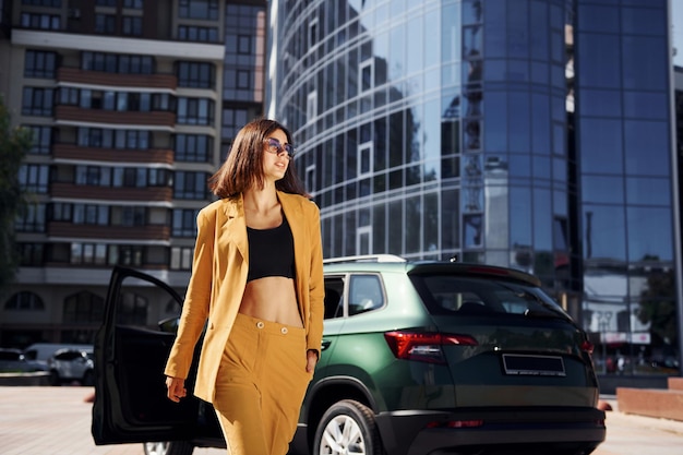 Walks forward from automobile Young fashionable woman in burgundy colored coat at daytime with her car