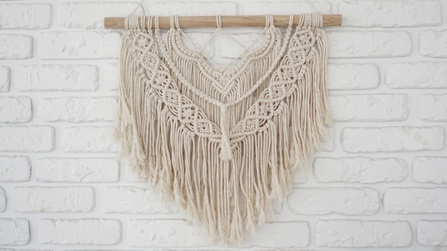 Wall panel in the style of Boho made of cotton threads in natural color on a white brick wall. Beautiful boho macrame wall panel for a cozy atmosphere