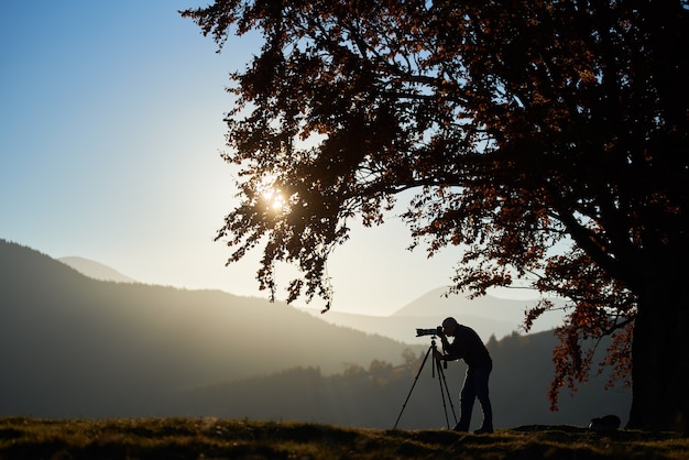 Wandelaar toeristische man met camera op met gras begroeide vallei op de achtergrond van berglandschap onder grote boom.