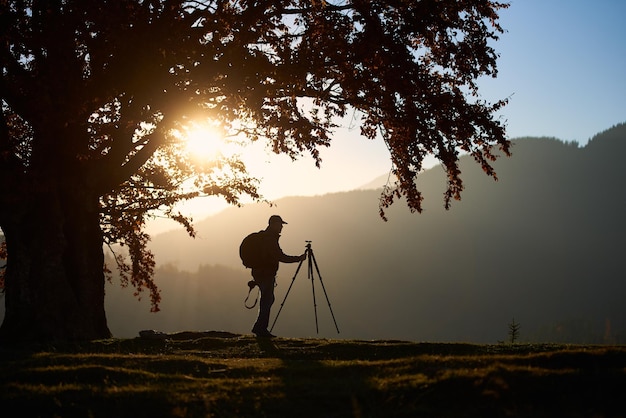 Wandelaar toeristische man met camera op met gras begroeide vallei op de achtergrond van berglandschap onder grote boom