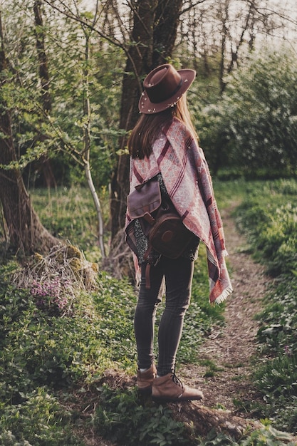 Wanderlust concept stylish hipster girl in hat with backpack walking in woods in evening sunshine woman traveler back view in sunlight among trees space for text atmospheric moment