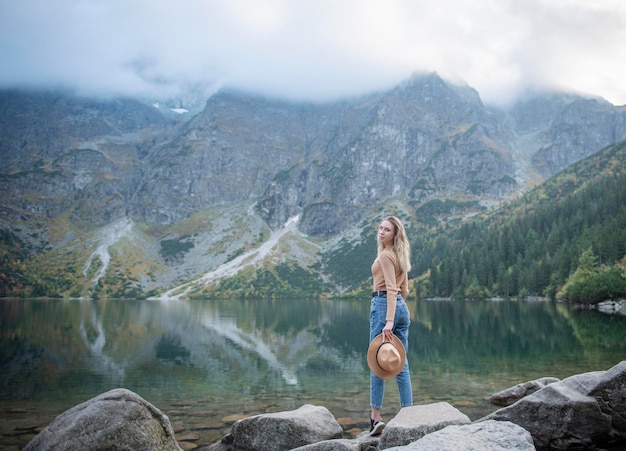 Wanderlust and travel concept. Stylish traveler girl in hat. Young woman with backpack travelling at lake in forest.