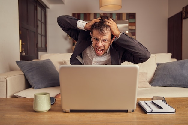 Wanhopige en boze jonge blanke man in pak zittend op een bank voor een laptop. Mislukking in bedrijfsconcept. Thuis op de bank zitten. Handen op het hoofd.