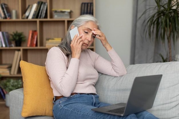 Wanhopige, vermoeide, kaukasische oude dame die op de bank zit, een telefoontje pleegt, naar de computer kijkt, lijdt aan