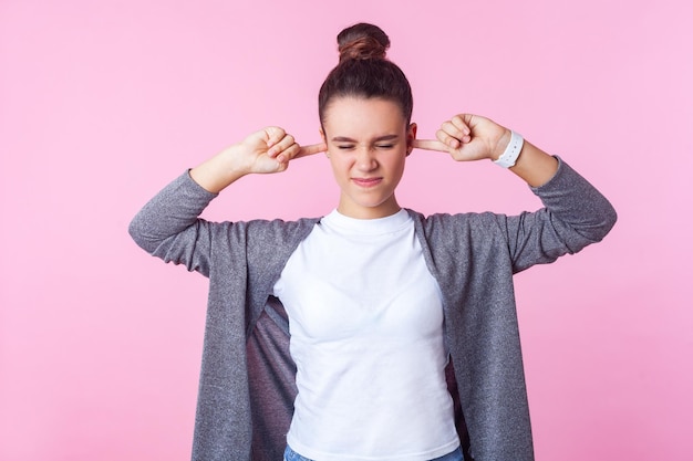 Don't want to listen. Portrait of naughty brunette girl with bun hairstyle in casual clothes covering ears ignoring parental upbringing, disobedient teenager. studio shot isolated on pink background