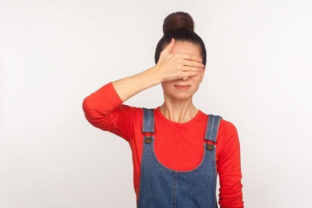 Don't want to look. Portrait of displeased girl with hair bun in denim overalls covering eyes, hiding grimace of disgust with hand, avoiding to watch. indoor studio shot isolated on white background