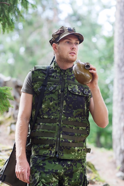 war, hiking, army and people concept - young soldier or ranger with gun drinking from flask in forest