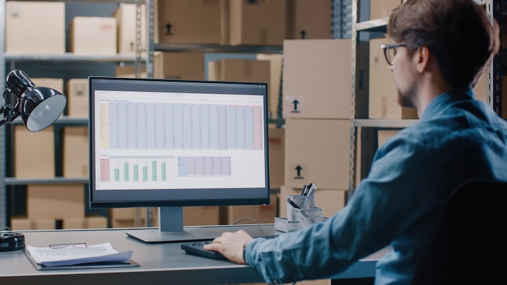 Photo warehouse inventory manager works with a spreadsheet on a personal computer while sitting at his desk in the background shelves full of cardboard box packages ready for shipping