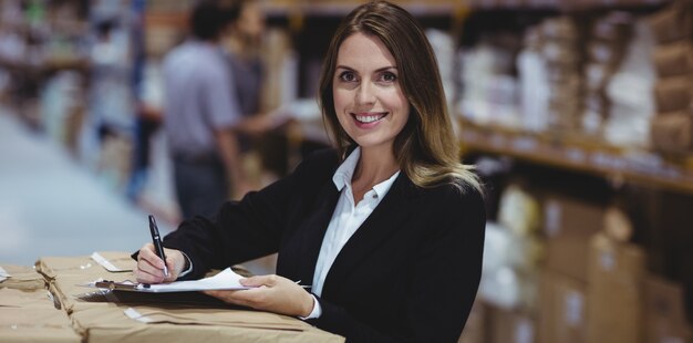 Warehouse manager writing on clipboard and smiling at the camera