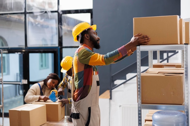 Warehouse package handler choosing cardboard box on shelf while standing on ladder. African american man storehouse worker preparing customer parcel and taking carton container