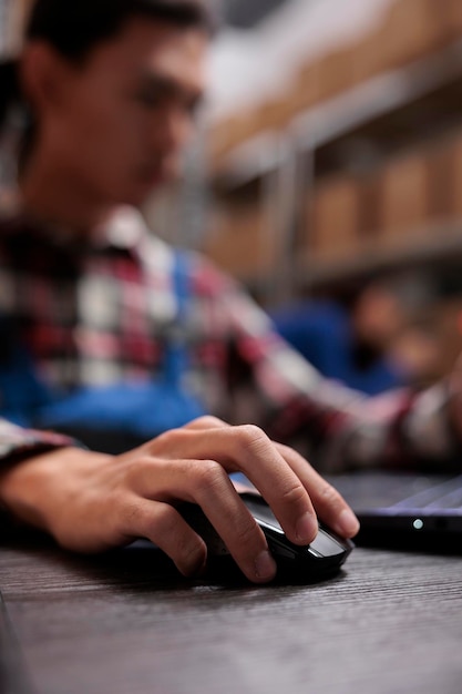 Warehouse worker creating parcel invoice on laptop in storage room. Storehouse employee sitting at desk, analyzing freight logistics report while using computer mouse close up