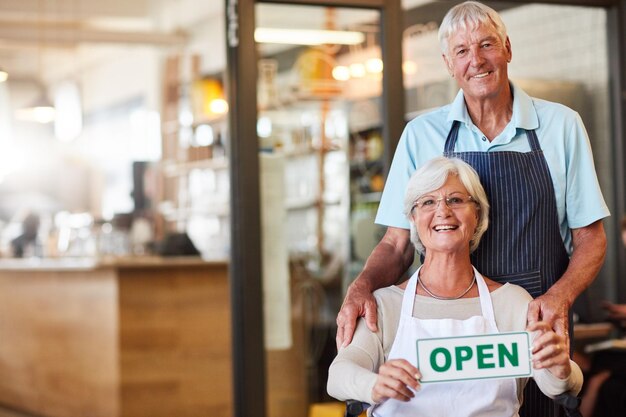 Waren ook open en rolstoelvriendelijk Portret van een gelukkig senior koppel met een open bord in hun winkel