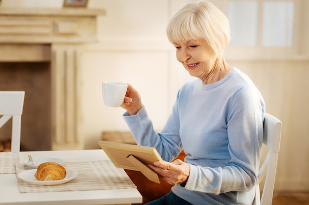 Warm atmosphere. Positive delighted blonde sitting in semi position and bowing head while drinking tea