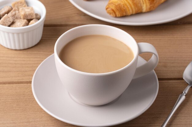 白いカップに温かいコーヒーを木製のテーブルでクロワッサンと砂糖の立方体で提供します