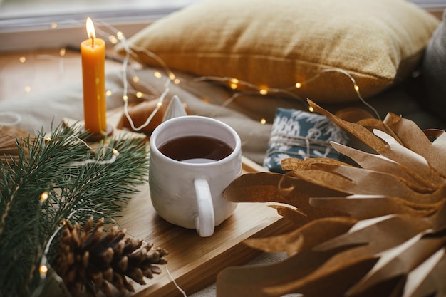 Warm cup of tea with christmas lights stars pine trees candle pillows on bed Cozy home hygge