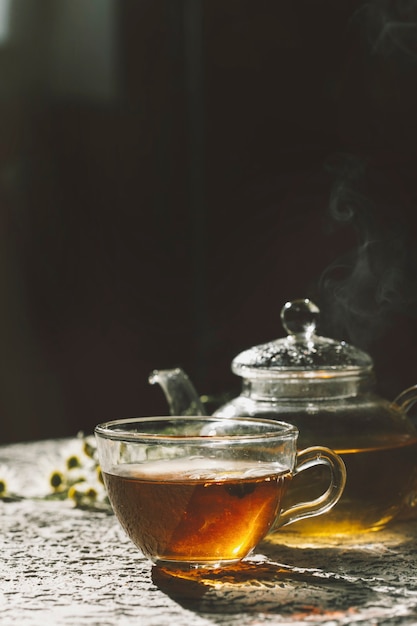 Warm cup of tea with glass teapot on dark background