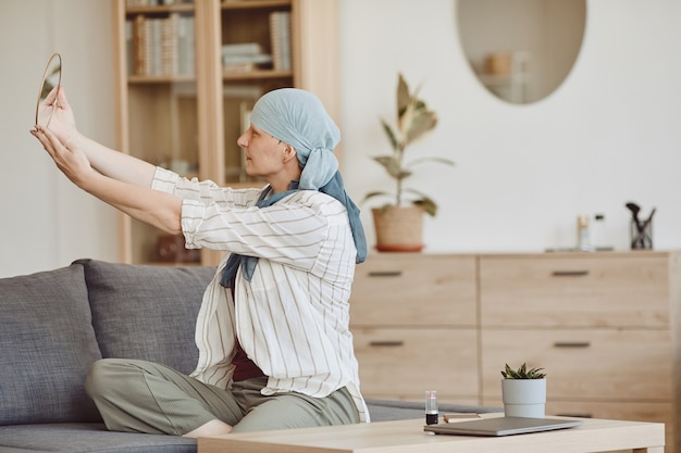 Warm-gestemde portret van kale vrouw die hoofddoek draagt die in spiegel kijkt terwijl het zitten op bank in gezellig huisbinnenland, exemplaarruimte