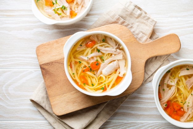 Теплый здоровый домашний куриный суп в белых керамических мисках на разделочной доске, белом фоне деревянного стола. традиционный вкусный куриный суп полезен для здоровья и иммунной системы. вид сверху
