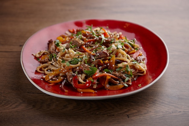 Warm noodle salad with meat and vegetables in a ceramic plate on a dark wooden table.