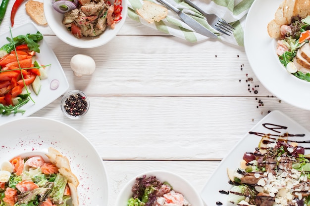 Warm salads frame on white wood flat lay. Top view on restaurant table with assortment of meat side