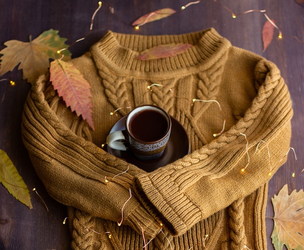 写真 暖かいセーターの袖は一杯のコーヒーを抱きしめ、秋の静物はコーヒーと黄色の葉と花輪で